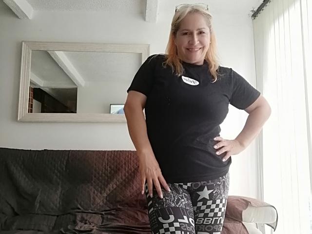 MajaReal stands smiling, blonde hair down, black t-shirt, checkered black and white pants, hands on hips, white-framed mirror behind, vertical blinds to the right.