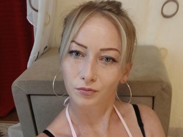 LeilahotX has blonde hair in a messy bun, blue eyes, pink lipstick, black halter top, large hoop earrings, and a slightly pouty face. She's standing in front of a grey box.