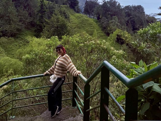 SaraleePoison leans on a green railing, wearing a striped sweater, black pants, and sunglasses, with a white crossbody bag, standing on a wooded hill.