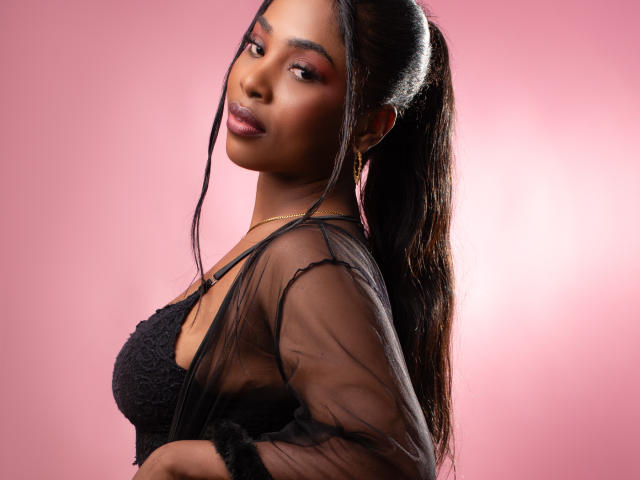 ZendayaRosse com pele escura, cabelo preto comprido em um rabo de cavalo, vestindo uma blusa preta transparente sobre um top de renda, sombra rosa nos olhos, olhando por cima do ombro, fundo rosa, sorriso discreto..