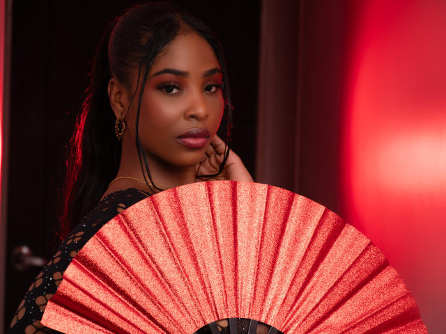 ZendayaRosse houdt een rode, glinsterende waaier vast en kijkt verleidelijk in de camera met een lichte glimlach. Ze heeft donker haar, bruine ogen en draagt gouden oorbellen..
