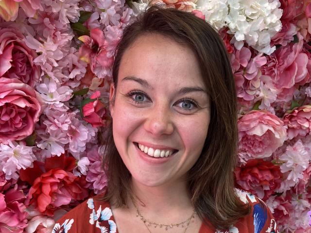 LibertineNila has cute blue eyes, a smiling face, and brown hair. She's wearing a floral dress, with pink and white flowers all around her, looking happy and pretty.