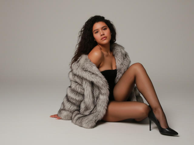 AriaVega with curly black hair, wearing a gray fur coat, black strapless top, sheer black stockings, and black high heels, sitting with legs crossed, showing her ass.