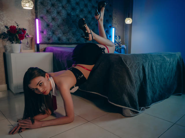 JennaDerose, con tacones negros y ropa interior roja, está acostada en el suelo de baldosas blancas, con las nalgas levantadas, debajo de una cama gris, con las piernas en el aire, mirando a la cámara, con cabello largo y oscuro, y un pañuelo blanco alrededor del cuello..