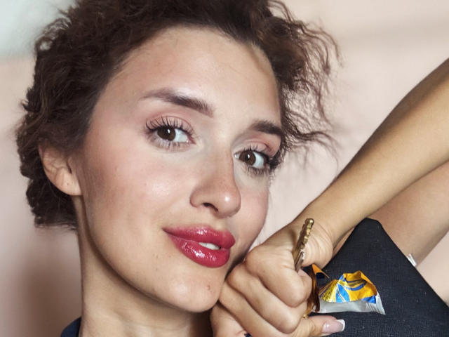 AnnaAngel heeft krullend bruin haar, glanzende roze lippen en een kleine gouden armband om haar rechterhand, terwijl ze haar kin op haar linkerpols rust. Ze glimlacht subtiel..