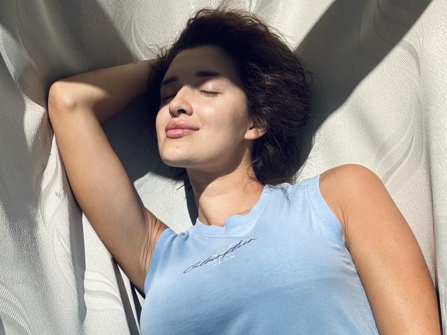 AnnaAngel com os olhos fechados, lábios rosa claro e cabelo escuro e desalinhado, vestindo uma blusa azul sem mangas, deitada com um braço atrás da cabeça, com a luz do sol no rosto..