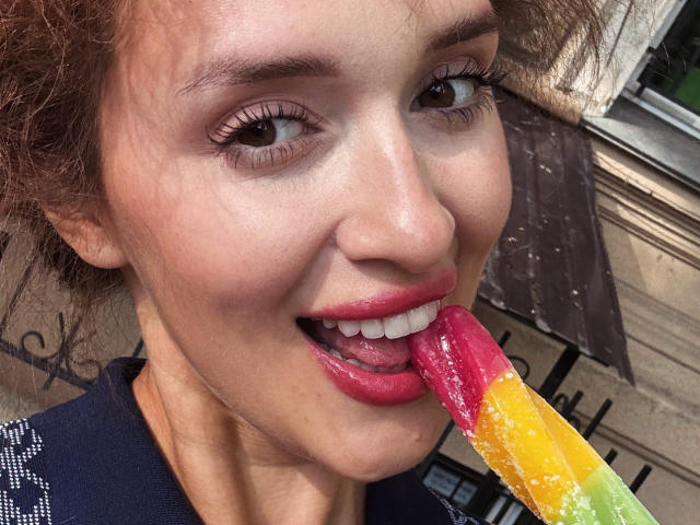AnnaAngel's face, mouth open, tongue out, licking a colorful, pink-to-yellow ice pop. She has curly brown hair, pink lipstick, and is wearing a dark blue shirt.