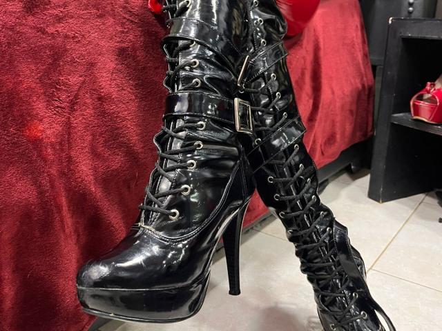 PaulaCamis' shiny black leather high-heel boots with lace-up front, buckle detail, and platform heel, standing on tiled floor, next to a red-covered surface.