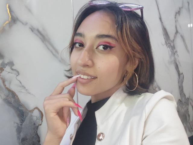 CamiVargas, com cabelo castanho, delineador rosa, brincos de argola, jaqueta branca, blusa preta, unhas rosa, dedo nos lábios, sorrindo, óculos de sol na cabeça, fundo de mármore..
