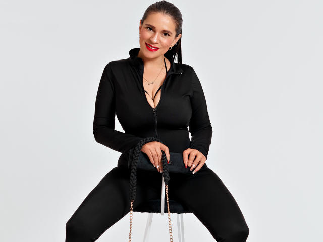 CelesteCorine con una treccia scura, una camicia nera con zip e pantaloni, seduta su uno sgabello, sorridente, rossetto rosso, sfondo bianco, mani che stringono la corda, seno di media grandezza, fisico atletico..