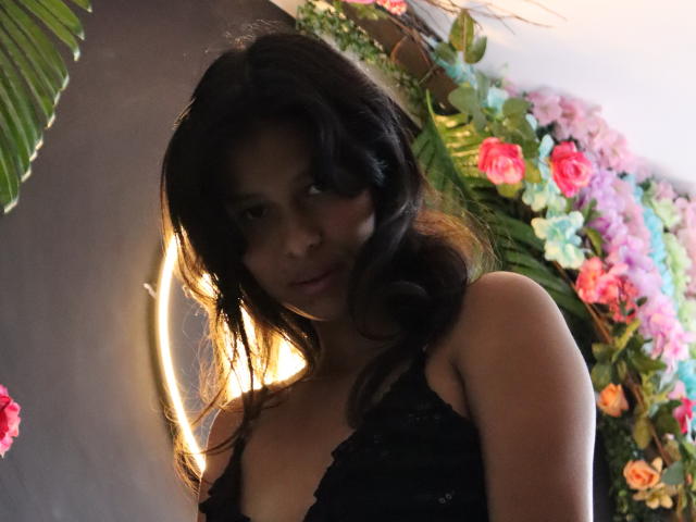AnickaTomzon, met donker golvend haar, een zwarte kanten top en een subtiele glimlach, staat in zacht licht, waarbij haar borsten zichtbaar zijn, voor een kleurrijke muur met bloemen..