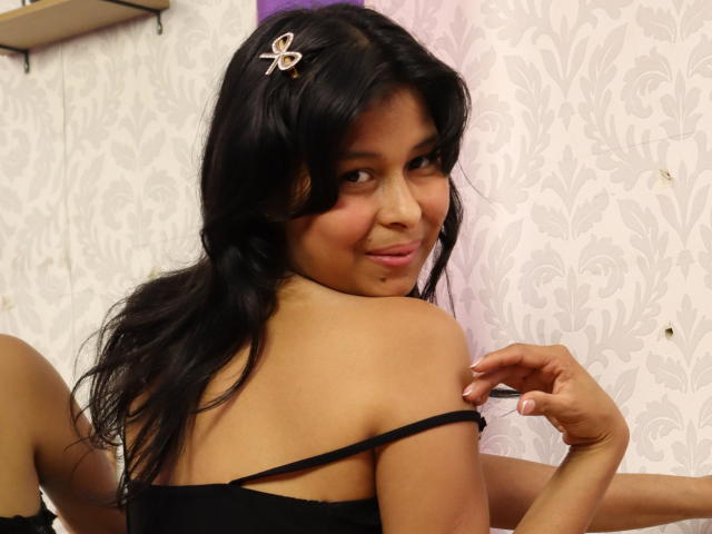 AnickaTomzon, avec ses cheveux foncés, une pince à cheveux en forme de papillon et une bretelle noire qui glisse de son épaule, sourit à l'appareil photo, montrant une petite tâche sur sa joue. Ses seins sont visibles..