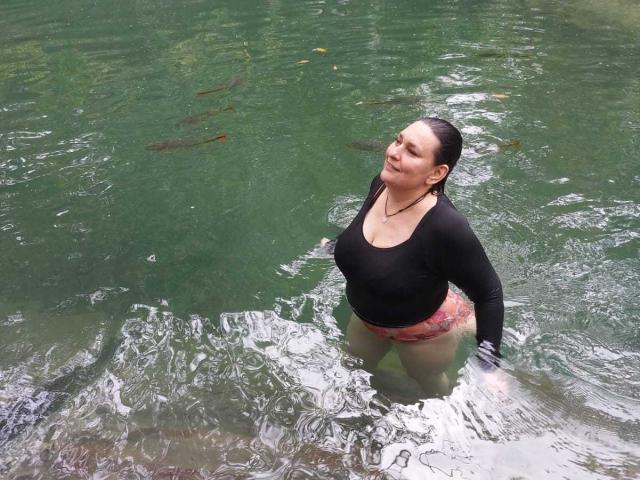 LadySelene standing in green water, wearing a black long-sleeve top and pink floral shorts, with her tits slightly visible, dark wet hair, eyes closed, smiling.