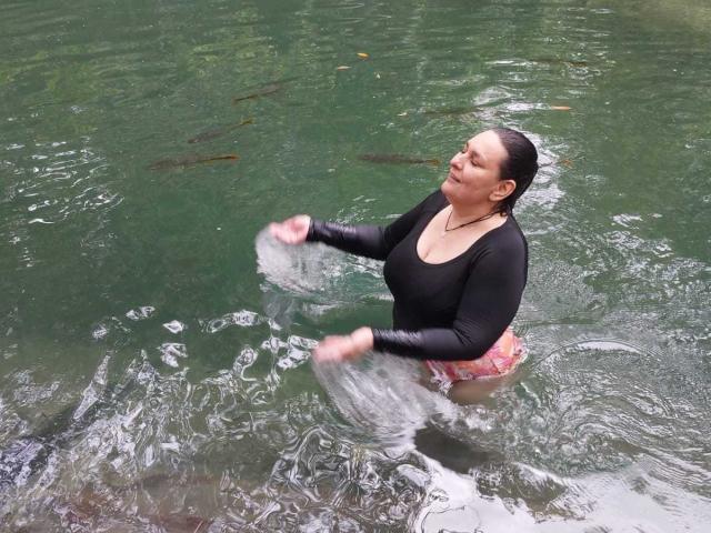 LadySelene has wet black hair, wearing a black long-sleeve top, and is splashing water with both hands, showing her pink and white patterned ass in the green water.
