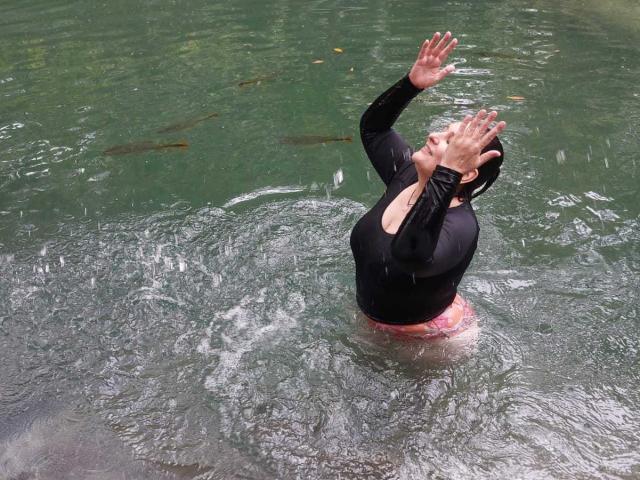 LadySelene with wet black top, raised hands, and pink bottomwear, standing in green water, looking up. Her tits are visible, and she has dark hair tied back.