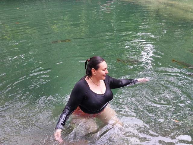 LadySelene with wet black top, large boobs, and red underwear, standing in green water, arms out, looking to the side, dark hair slicked back.
