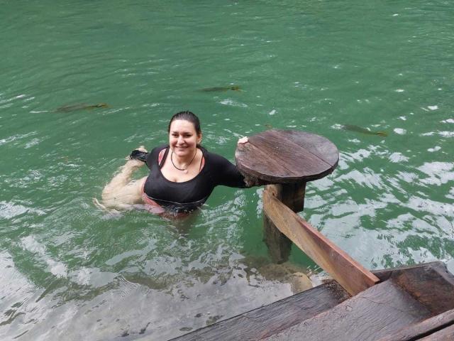 LadySelene with wet black shirt and red shorts, smiling, standing in green water, leaning on wooden dock, right arm on round table, water up to her thighs.