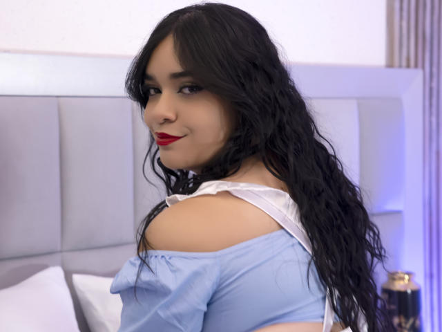 AliciaRelas with long black wavy hair, red lipstick, and a blue off-shoulder top, looking back over her shoulder, showing her bare shoulders and white lace collar.