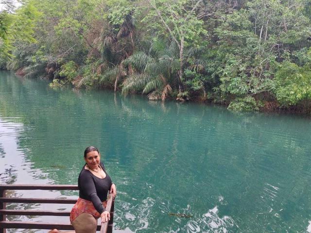 LadySelene with brown skin, black long-sleeve top, and orange skirt, sitting on wooden stairs by a clear green river, forest in background, smiling at camera.