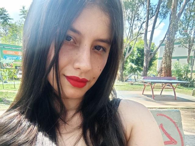 MadissonFontaine with dark, straight hair, red lipstick, and freckles, smiling at the camera, showing her bare shoulders. Her tits are not visible. She's in a sunny park.