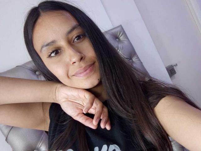 LizyGrey with long black hair, brown eyes, and a small mole on her chin, smiling slightly, resting her head on her hand, wearing a black shirt, in a silver chair.