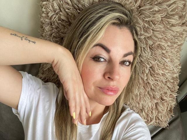DanniaMoore with blonde hair, pink lips, and yellow nails, lying on a fluffy beige pillow, wearing a white shirt, and sporting a tattoo on her left forearm.