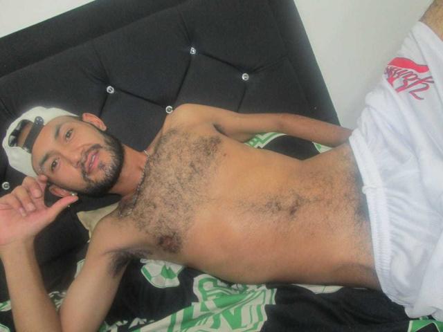 NatashaAndKrillyn, shirtless with a hairy chest, lying on a bed, wearing white shorts, and a white cap, has a beard and is looking at the camera.
