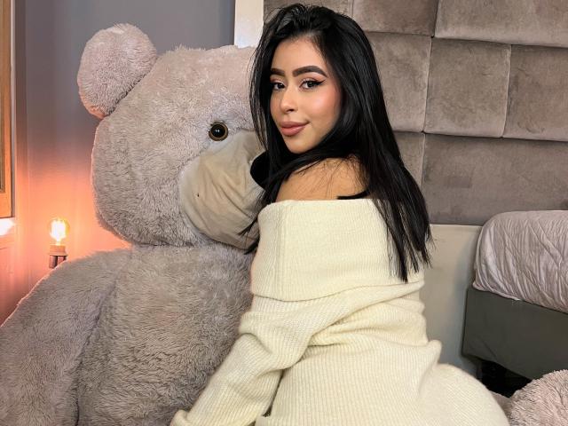 MilaGonzalez with long black hair, off-shoulder white knit sweater, sitting next to giant grey teddy bear, looking over shoulder with subtle smile, bedroom setting.