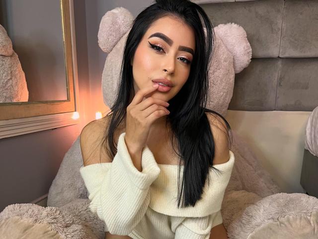 MilaGonzalez with long black hair, wearing an off-shoulder white sweater, touching her lips, sitting in front of a large beige teddy bear, with soft pink lighting.