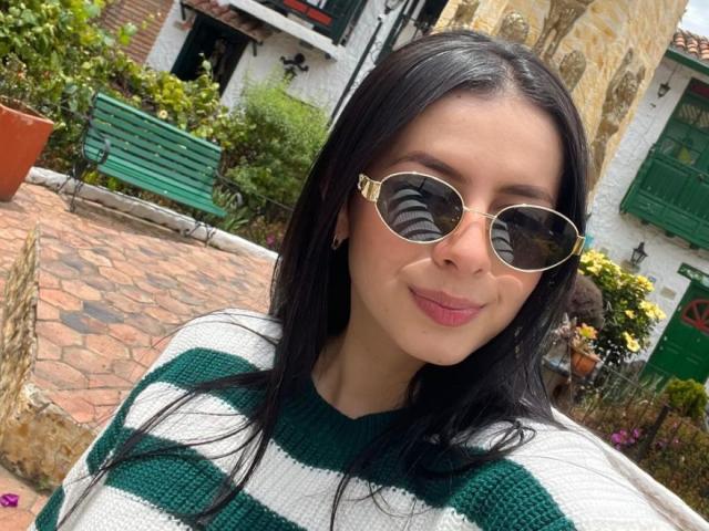 Isabella James tem cabelos longos e pretos, veste um suéter listrado de verde e branco, e usa óculos de sol redondos dourados com lentes refletivas, sorrindo com batom rosa..