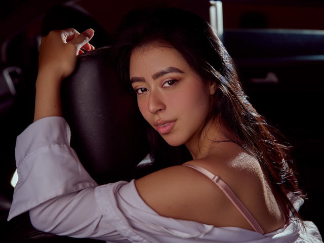 MilaGonzalez with dark hair, tan skin, and pink bra strap, wearing a white shirt, leaning on a black car seat, looking back with a slight smile.