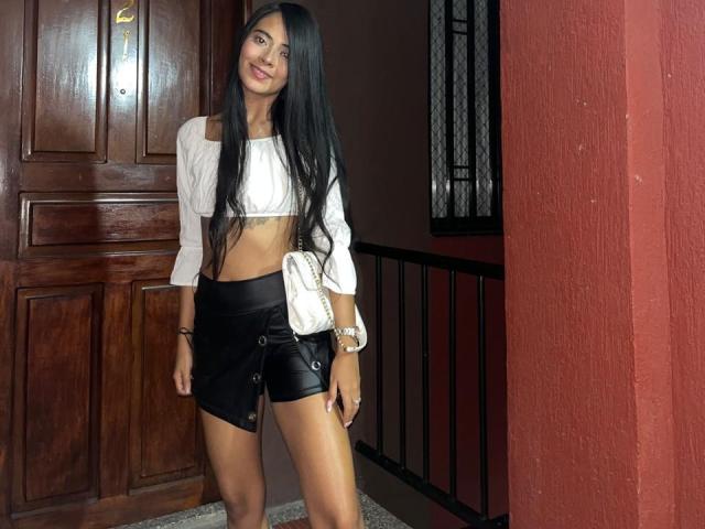 TamaraPeach, com cabelos longos e pretos, blusa cropped branca com um ombro à mostra, shorts de couro preto, bolsa branca, sorrindo, em pé na porta, porta marrom, parede vermelha, corrimão preto..