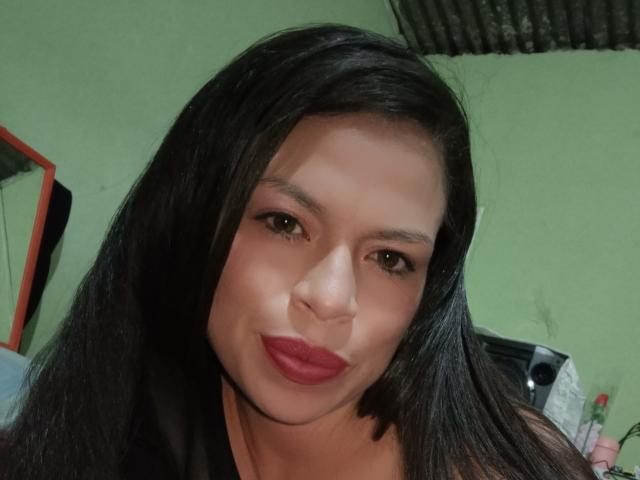SharydHot tem cabelos pretos longos, olhos castanhos e batom vermelho, fazendo uma expressão de lábios franzidos com as sobrancelhas levemente arqueadas. Ela está vestindo uma blusa preta e parece adorável..