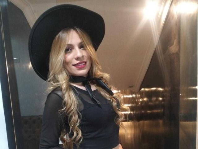 AmbarVelle has long blonde hair, wears a black hat and sheer top with a black ribbon, smiling with red lipstick, showing her tits under the transparent top.