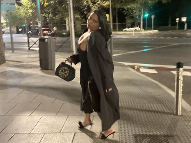 Chili walks street at night, wearing black outfit, holding black purse, long black hair, city lights in background, streetlamp.