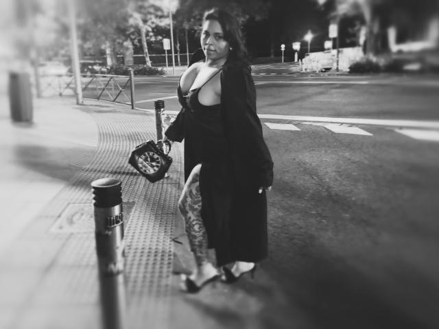Chili è in piedi su un marciapiede della città in bianco e nero, indossando un abito con un enorme decoltè, tenendo una borsa.