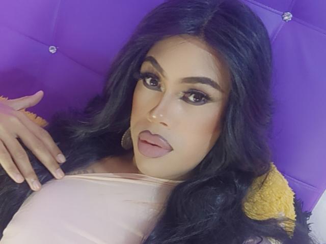 CrystalNew has long black hair, thick eyebrows, and full lips with pink lipstick. She's lying on a purple surface, tongue slightly out, wearing a pale pink top.