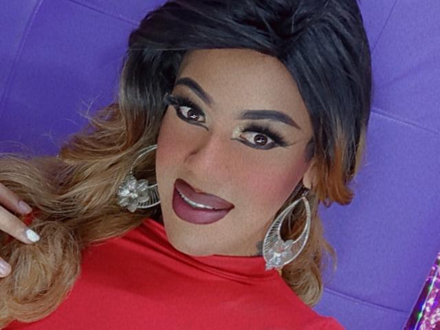 CrystalNew with dark brown, wavy hair, thick eyebrows, and red lipstick, wearing a red turtleneck, large silver earrings, and a confident smile, showing off her pouty lips.
