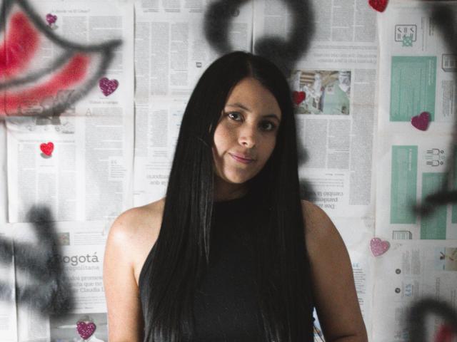 EmilySavage with long black hair, wearing a black sleeveless top, standing in front of a wall covered in newspaper, small hearts, and graffiti. She has a slight smile.