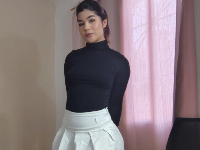 ScarlletZhen, wearing a black turtleneck that shows her small tits and a white pleated skirt, stands with arms behind her back, pink curtains in the background.