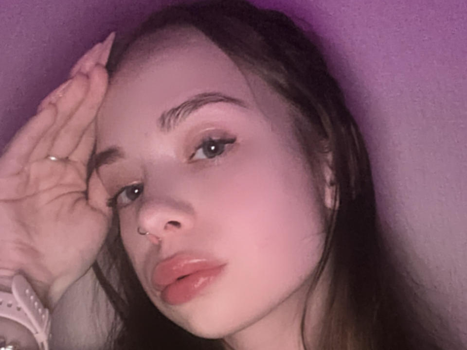 CelieBrooks with long brown hair, full pink lips, and a nose ring, touching her head with a hand, wearing a white bracelet, against a purple background.