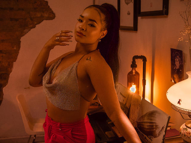 MilaRomano with tan skin, black ponytail, small tattoos, silver mesh top, revealing side, red pants, hand near face, warm orange lighting, lantern, chair, and lamp in background.