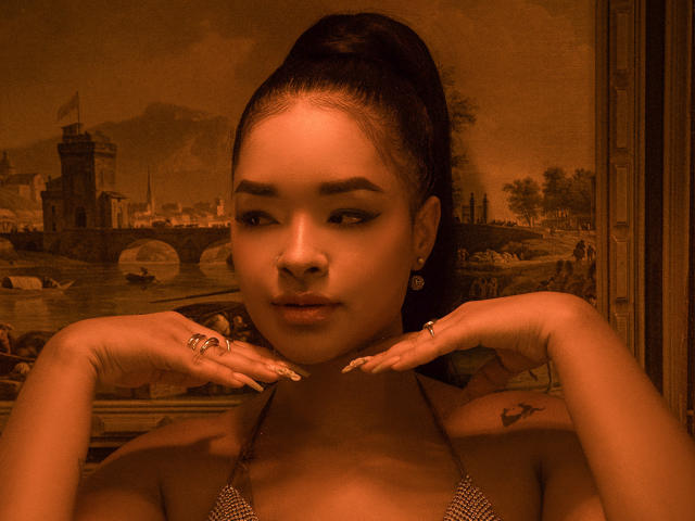 MilaRomano with dark hair in a ponytail, wearing a sparkly top, hands near her neck, wearing rings and earrings, brown-tinted image, small heart tattoo on her right shoulder.