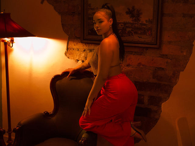 MilaRomano with long black hair in a ponytail, wearing a red skirt, exposing her ass, and a lace bra, leaning on a dark chair, under warm lamp light.