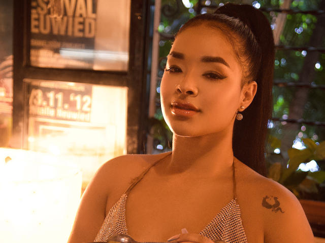 MilaRomano has a pretty face, dark ponytail, shiny brown skin, small tits in a sparkly halter top, nose ring, tattoo on right shoulder, and earrings, looking at camera.