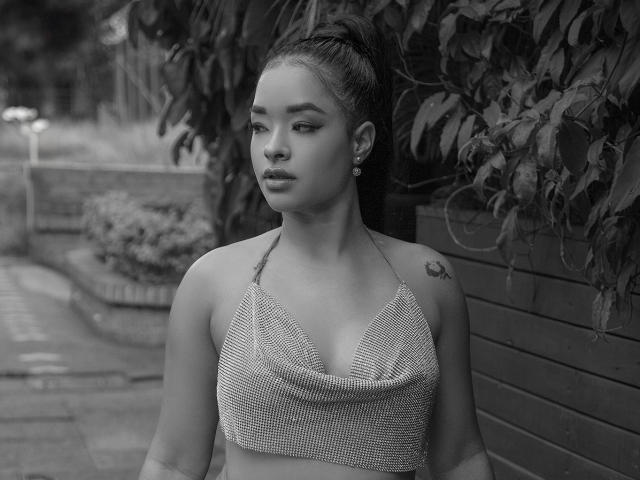 Black-and-white photo of MilaRomano with medium tits, wearing a checkered halter top, hair in a high ponytail, looking to the side, small tattoo on right shoulder, earrings.