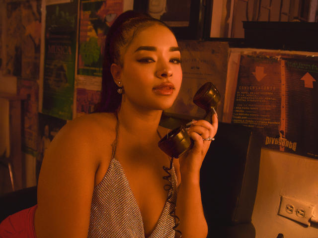 MilaRomano with brown skin and purple hair holds a vintage brown phone, wearing a checkered top with a low neckline, showing her shiny lips and small earrings.