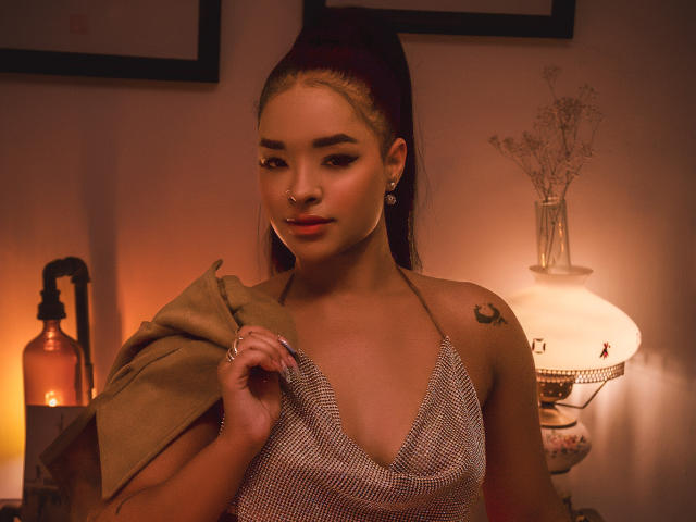 MilaRomano with dark ponytail, nose ring, and earrings, wearing a mesh halter top, holding a tan coat, soft orange lighting, visible tattoo on right shoulder.