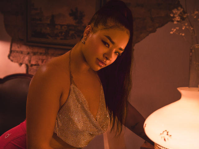 MilaRomano with dark hair in a high pony, wearing a sparkly mesh bralette, pink pants, and ear studs, looking down with a soft, seductive smile, near a warm lamp.