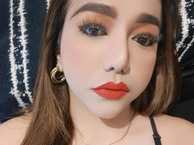 FabulousNina69 has red lipstick, brown eyes with long eyelashes, brown eyebrows, gold hoop earrings, and brown hair. She's wearing a black strap top.